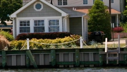 A quaint house by the water, surrounded by greenery and a wooden dock, with a grill visible in the background.