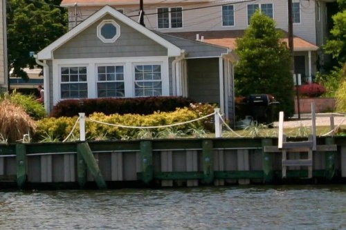 A waterfront view of a gray house with large windows, surrounded by greenery and a wooden dock.