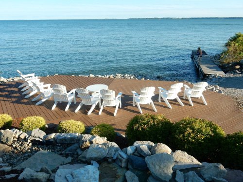 A serene lakeside scene with white chairs arranged on a deck, overlooking calm water and a distant shoreline.