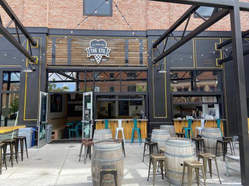 Exterior of a bar called "The Still," featuring wooden barrels, outdoor seating, and a rustic brick backdrop.