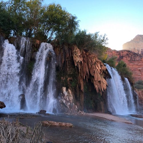 A scenic view of cascading waterfalls surrounded by lush greenery and rocky cliffs under a clear blue sky.