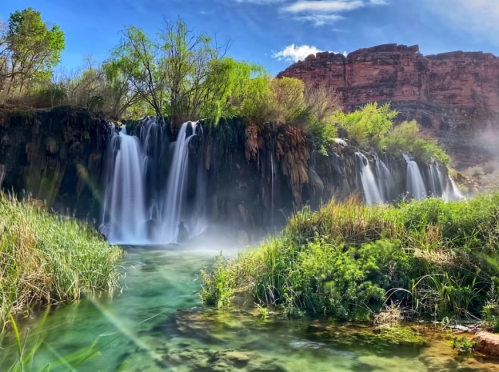 A serene waterfall cascades into a clear pool, surrounded by lush greenery and rocky cliffs under a blue sky.