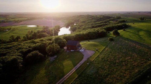 Aerial view of a house surrounded by lush greenery and a serene pond at sunset.