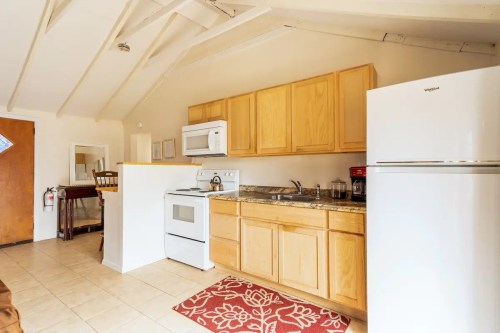 Bright kitchen with wooden cabinets, white appliances, and a red rug, featuring a dining area in the background.