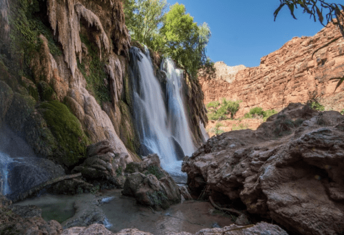A serene waterfall cascades over rocky cliffs, surrounded by lush greenery and red rock formations under a clear blue sky.