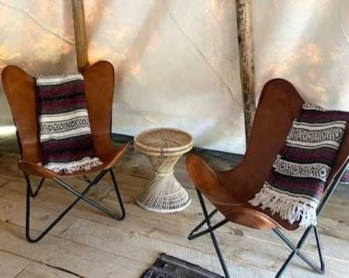 Two leather butterfly chairs with patterned blankets, beside a woven side table, set in a cozy, rustic interior.