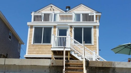 A two-story beach house with large windows, a front porch, and stairs leading up to it, set against a clear blue sky.