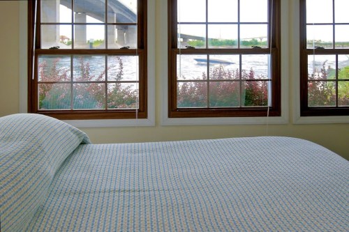 A cozy bedroom with a blue patterned bedspread, featuring large windows overlooking a river and a bridge.