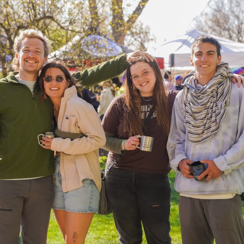 A group of four friends smiles together outdoors, holding mugs, with tents and trees in the background.