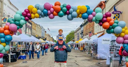 A man carries a boy on his shoulders under a colorful balloon arch at a bustling street market.