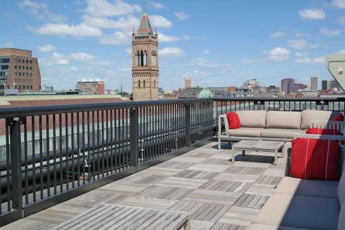 Rooftop terrace with modern seating, overlooking a city skyline and a clock tower under a blue sky with clouds.