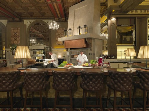 Two chefs prepare food in an elegant restaurant kitchen with a large fireplace and stylish seating.