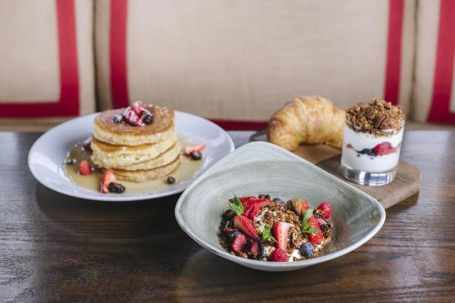 A plate of pancakes with berries, a bowl of granola with yogurt and fruit, and a croissant on a wooden table.