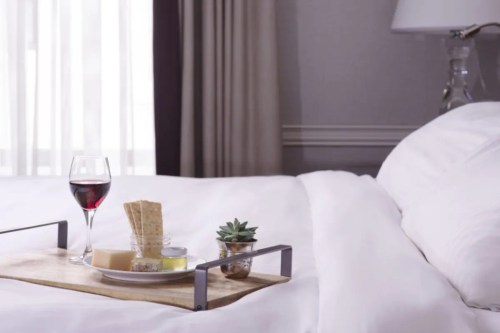 A cozy bed with a tray featuring wine, crackers, cheese, and a small plant, set against a soft, light backdrop.