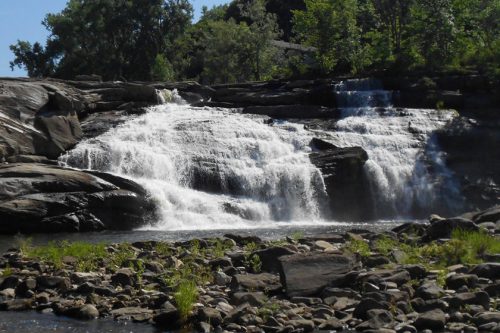 A cascading waterfall flows over rocky terrain, surrounded by lush greenery and a clear blue sky.