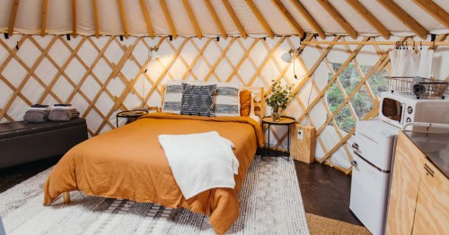 Cozy yurt interior with a bed, decorative pillows, and a small kitchenette, featuring wooden walls and warm lighting.