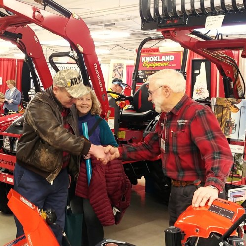 Two people shake hands at a trade show, surrounded by agricultural equipment and displays. A third person smiles nearby.