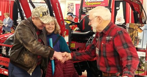 A man in a cap shakes hands with another man while a woman smiles nearby, all in front of red tractors at an event.