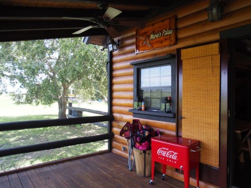 A rustic wooden porch with a Coca-Cola cooler, golf clubs, and a sign reading "Mayer's Palace" near a tree.