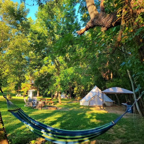 A peaceful outdoor scene featuring a hammock, tents, and lush greenery under a clear blue sky.