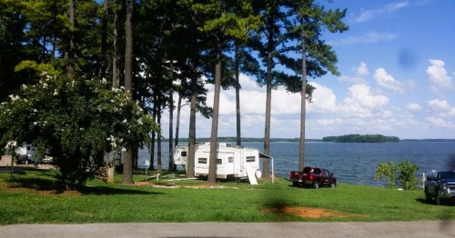 A serene lakeside scene with a camper, trees, and parked vehicles under a blue sky with fluffy clouds.