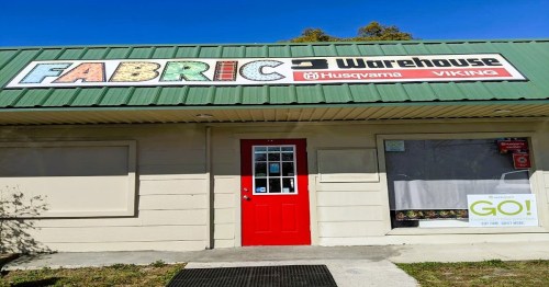 Exterior of a fabric warehouse with a green roof, colorful sign, and a red door. Large windows display a sign for a promotion.