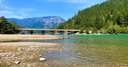 A serene river scene with a bridge, surrounded by lush green mountains and a clear blue sky.