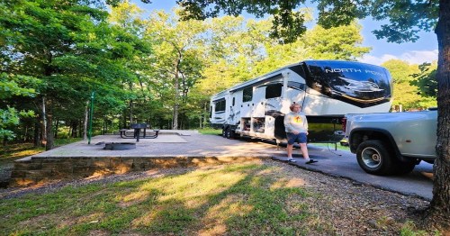 A man stands next to a large RV parked in a wooded area, with a picnic table and fire pit nearby.