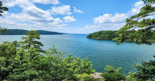 A serene lake surrounded by lush greenery and distant hills under a partly cloudy sky.