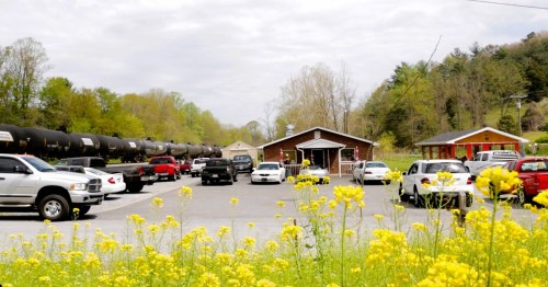 A parking lot filled with cars, surrounded by yellow flowers, with buildings and a train in the background.