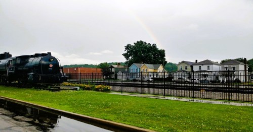 A vintage steam locomotive beside a grassy area, with colorful houses and a rainbow in the background under a cloudy sky.
