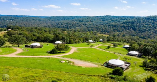 Aerial view of a green landscape with several yurts and winding paths surrounded by trees and hills.