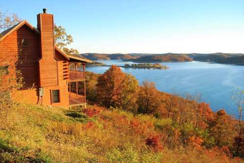 A wooden cabin overlooks a serene lake surrounded by vibrant autumn foliage and rolling hills.