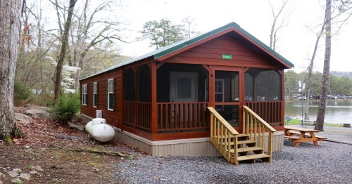 A cozy wooden cabin with a porch, surrounded by trees and near a calm lake.