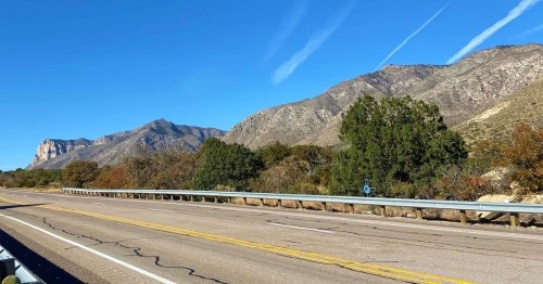 A clear blue sky over a highway with mountains and greenery on the side, showcasing a scenic landscape.