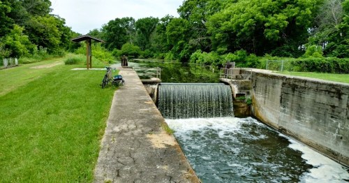 A serene waterway with a small waterfall, grassy banks, and a bench nearby under a cloudy sky.