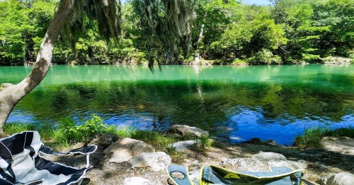 A serene river scene with green water, surrounded by lush trees and rocks, featuring a few chairs by the shore.