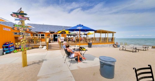 A beachside restaurant with colorful signs, outdoor seating, and umbrellas, overlooking the water on a sunny day.