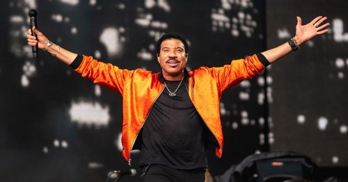 A performer in an orange jacket raises arms on stage, smiling and holding a microphone, with a blurred background.