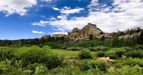 A scenic landscape featuring rocky formations, green foliage, and two people exploring near a stream under a blue sky.