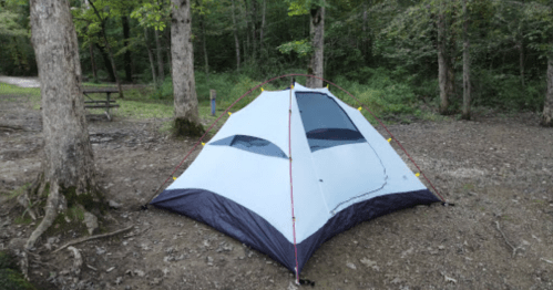 A camping tent set up in a wooded area, surrounded by trees and a gravel ground.