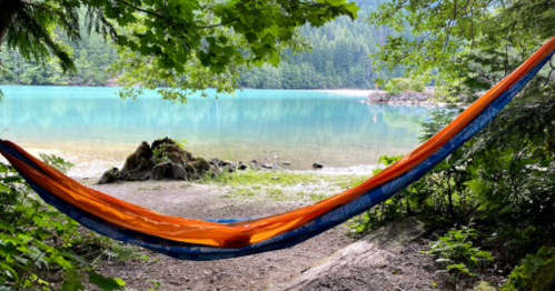 A vibrant orange and blue hammock hangs between trees by a serene turquoise lake surrounded by lush greenery.