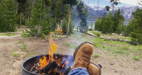 A pair of boots resting on a fire pit with flames, surrounded by trees and mountains by a lake in the background.