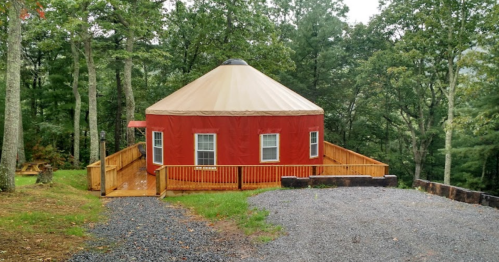 A red yurt with a tan roof, surrounded by trees, featuring a wooden deck and gravel pathway.