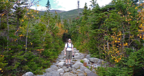 A hiker stands on a rocky trail surrounded by lush green trees and autumn foliage in a mountainous area.