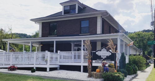 A brick restaurant with a white porch and sign, surrounded by flowers and decorative pumpkins, on a sunny day.