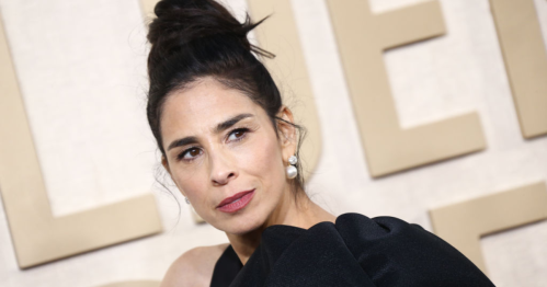 A woman with a bun hairstyle and pearl earrings poses in a black dress against a neutral background.