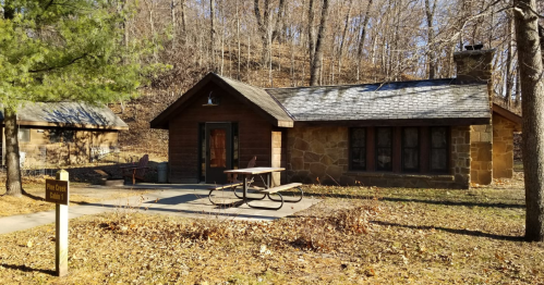 A rustic stone cabin with a picnic table outside, surrounded by trees and a wooded hillside.