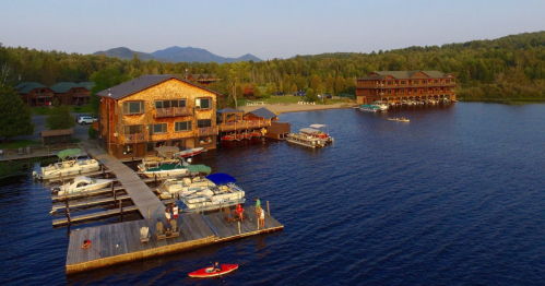 A scenic lakeside view featuring wooden buildings, boats docked, and people enjoying the water under a clear sky.