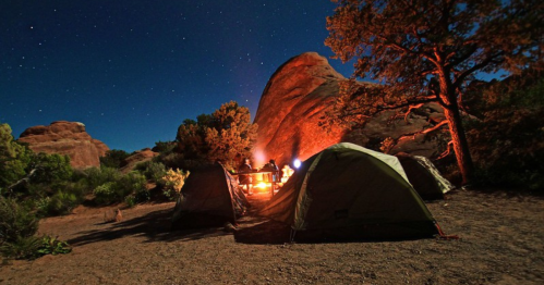 A campsite at night with tents, a glowing campfire, and rocky formations under a starry sky.